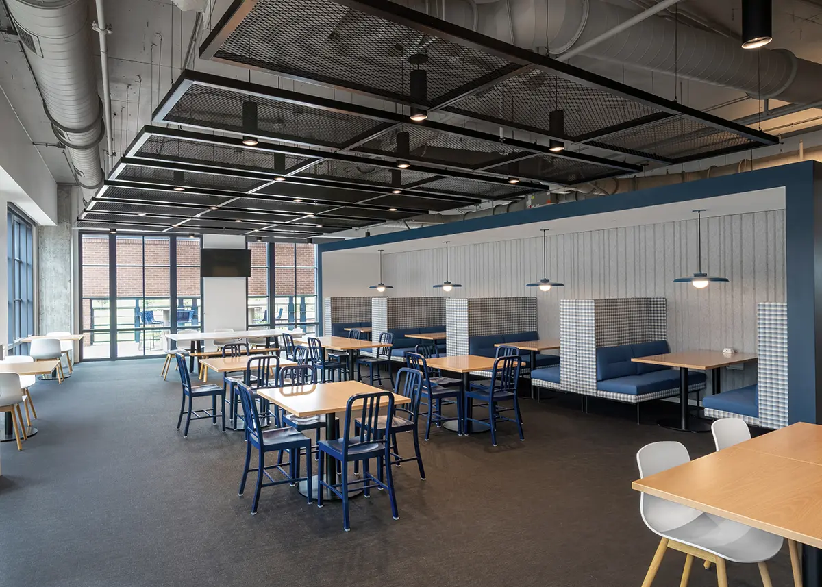 Spacious cafeteria with modern design, featuring blue and wooden booths, pendant lights, and large windows. The atmosphere is clean and inviting.