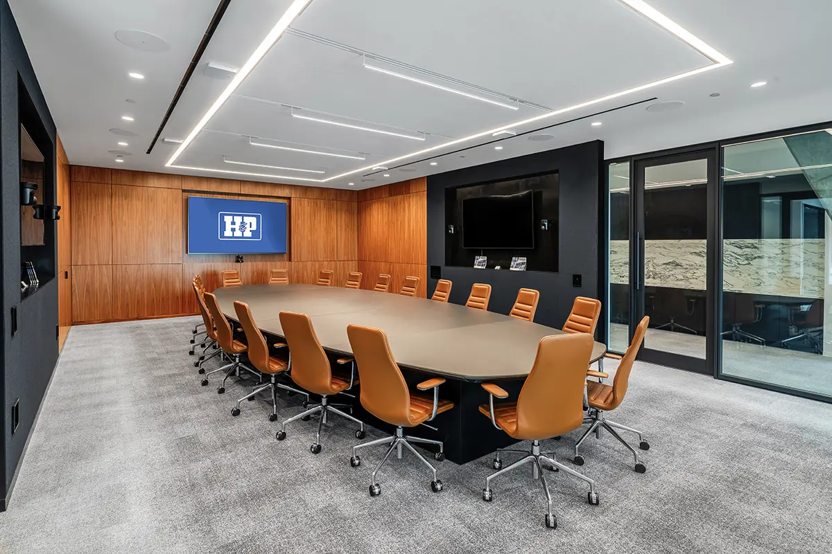 Spacious conference room with an oval table, surrounded by orange chairs. A screen on the wood-paneled wall displays a logo. Bright, modern lighting.