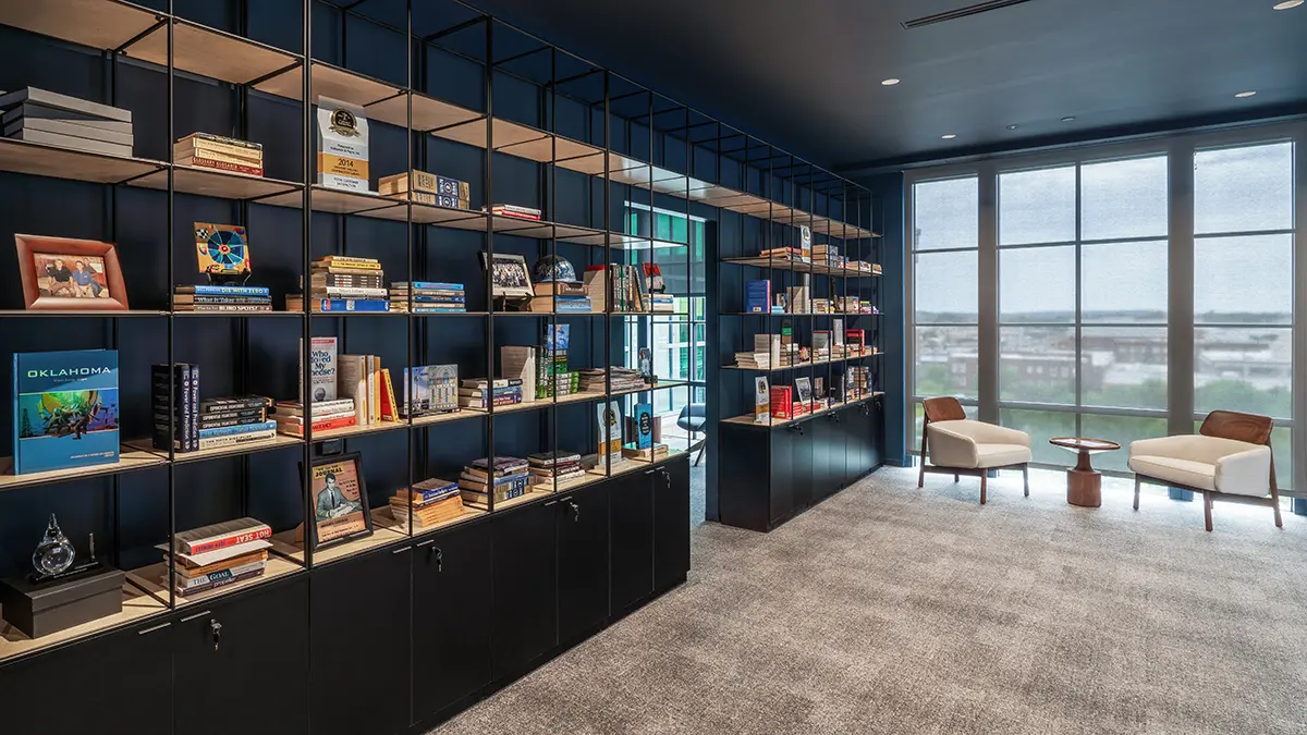 Modern library with sleek black shelving filled with colorful books, large windows allowing natural light, and two cozy chairs with a small table, creating a calm and inviting atmosphere.