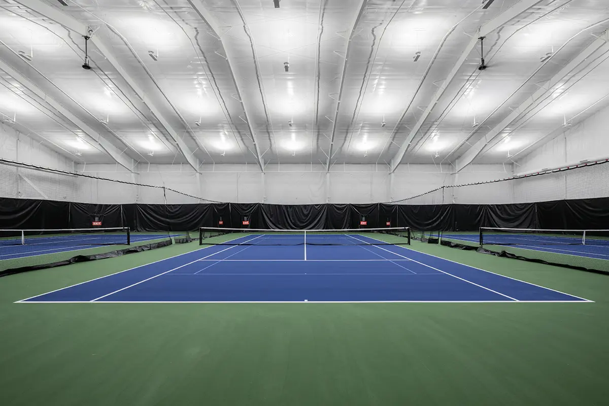 Indoor tennis court with green flooring and bright blue playing surface. Overhead white lights illuminate the spacious, high-ceilinged facility. Quiet and serene atmosphere.