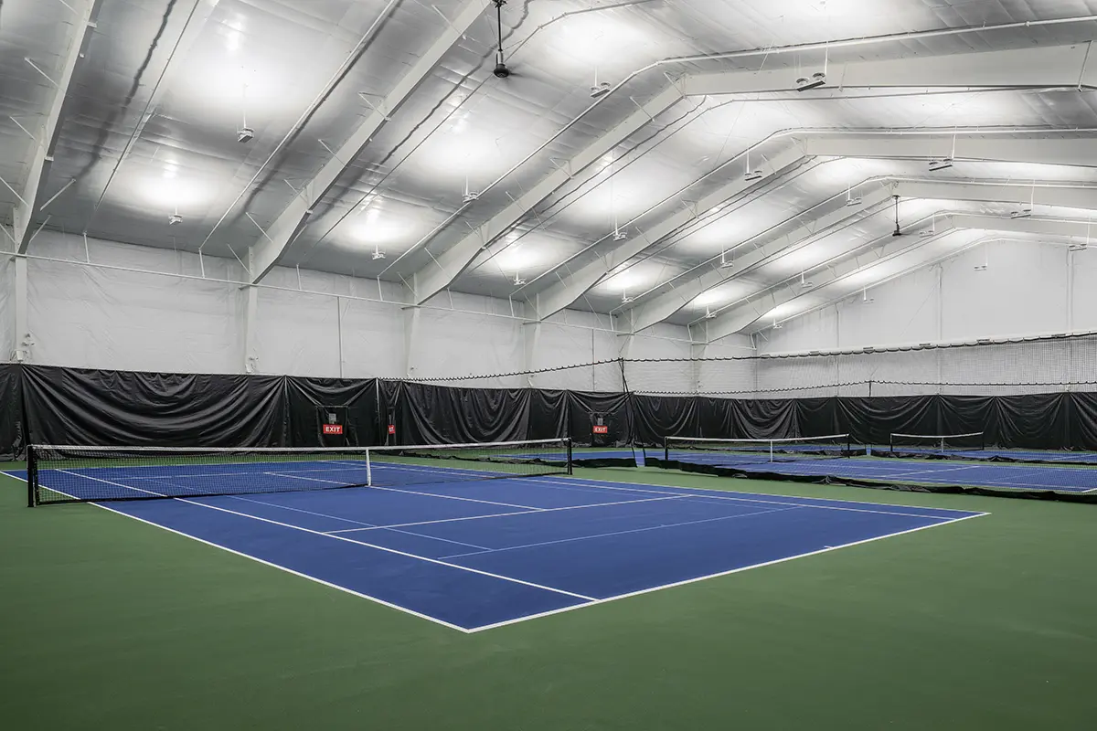 Indoor tennis court with a blue playing surface and green surrounding area. High ceiling with bright lights; nets divide multiple courts. Calm and orderly atmosphere.