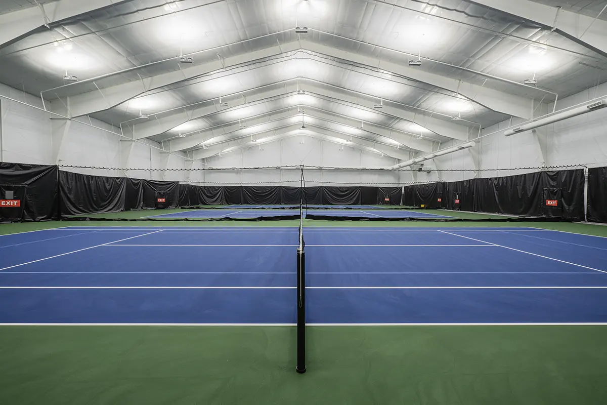 Indoor tennis court with blue-green flooring, lined by black curtains and marked exits. Bright overhead lighting creates a clean, modern ambiance.