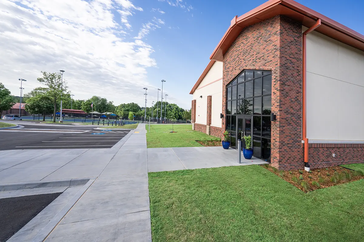 A modern brick building with large glass doors sits beside a tidy lawn under a bright blue sky. The adjacent parking lot is mostly empty.
