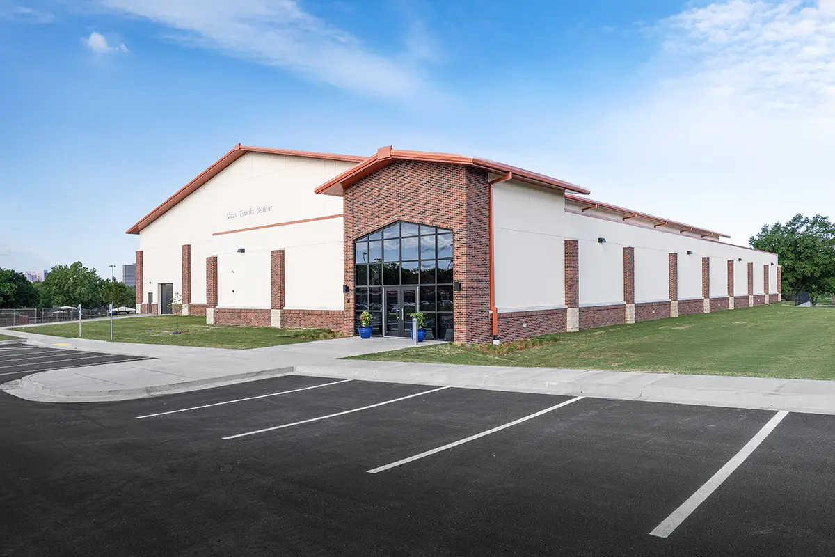 Large community center with a brick facade and white walls, under a clear blue sky. The building has a spacious entrance and an empty parking lot.