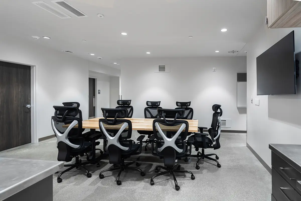 Modern conference room with a long wooden table and eight ergonomic chairs. White walls, mounted TV, and bright LED ceiling lights create a clean, professional atmosphere.