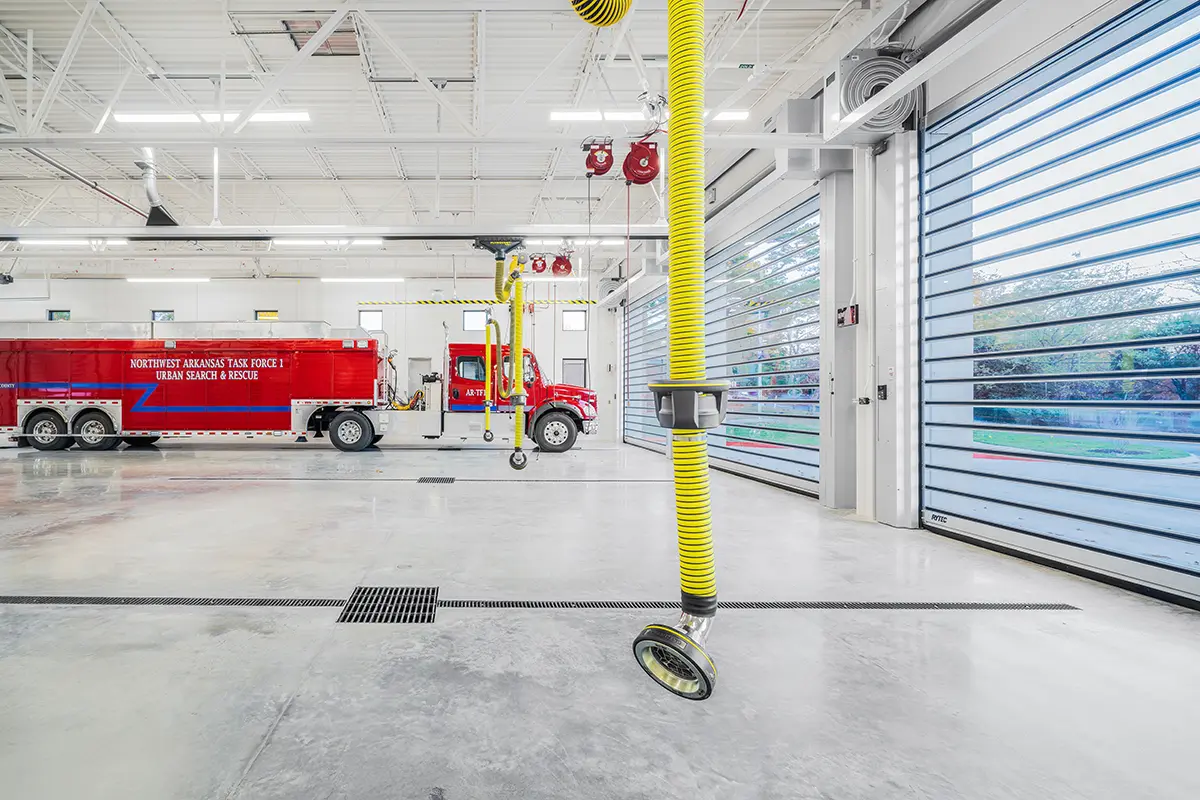 Interior of a fire station with a red Urban Search and Rescue truck, bright yellow exhaust hoses, and large blue garage doors. Clean and organized.