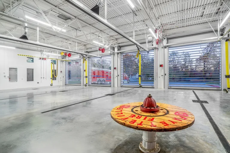 A spacious, modern fire station interior with bright lighting and garage doors open, revealing a red fire truck outside. A fire hydrant is mounted on a wooden spool table in the foreground. The space is clean and organized, conveying readiness.