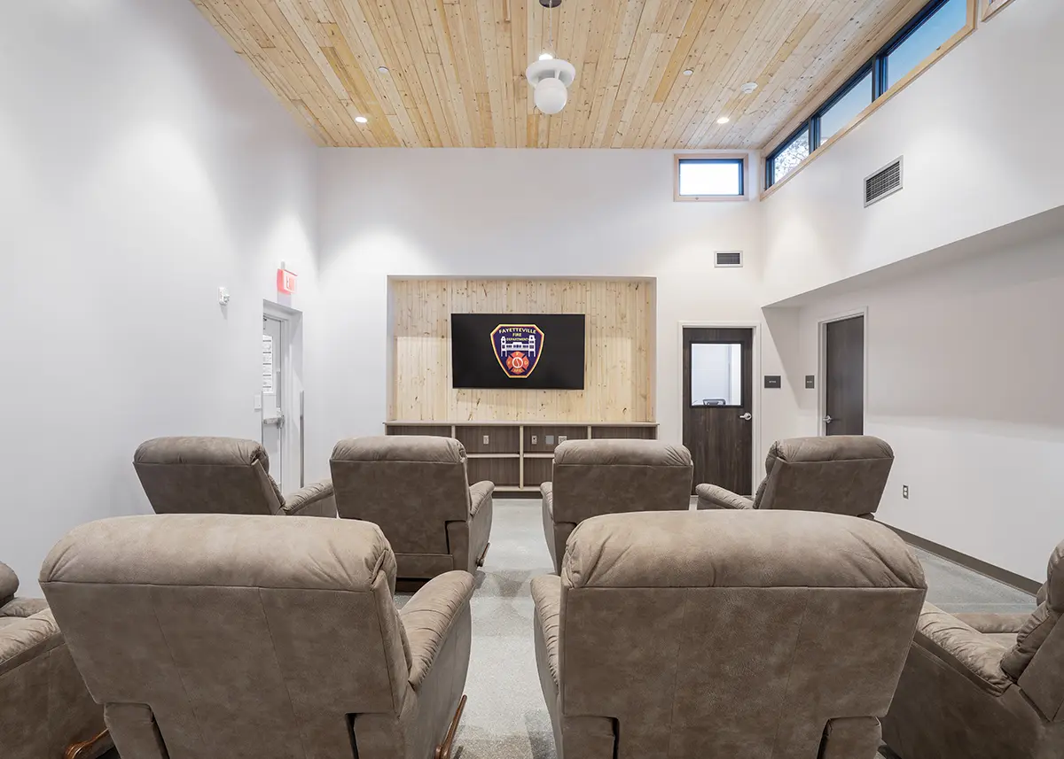 Cozy room with beige recliners facing a wooden wall-mounted TV displaying a fire department emblem. Ceiling features a warm, light wood finish and small windows.