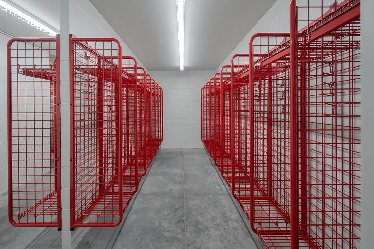A room with rows of empty red metal lockers on both sides, set against plain white walls. The lighting is bright and the floor is smooth concrete.