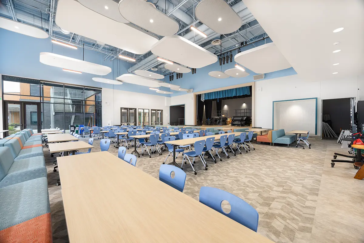 Spacious, modern cafeteria with blue chairs, light wood tables, stage, cushioned benches, and hanging ceiling panels. Bright, airy atmosphere.