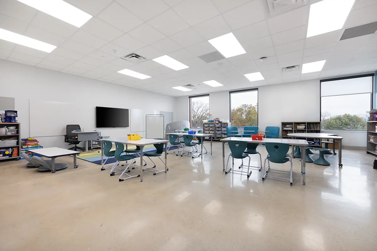 Modern classroom with white walls and large windows, filled with blue and white desks and chairs. Shelves hold supplies; a calm, organized atmosphere.