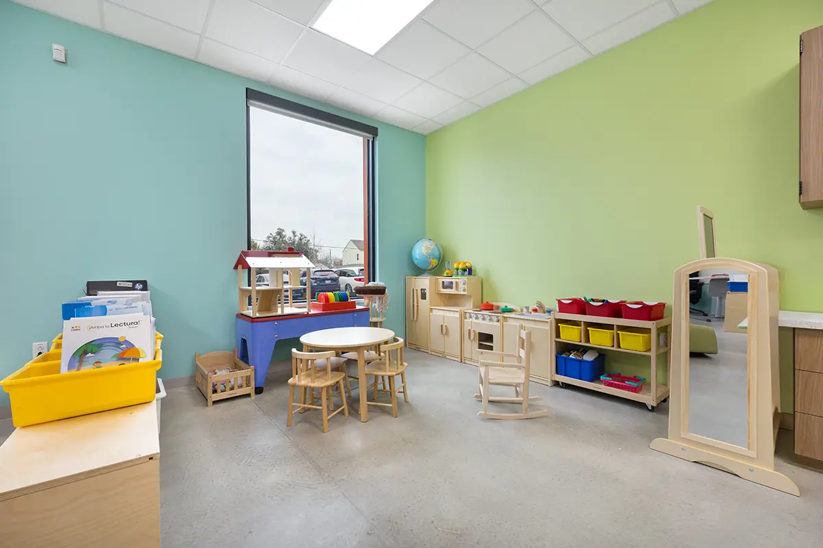 A bright, tidy playroom with pastel walls, a large window, round table with four wooden stools, toy kitchen set, colorful bins, and a globe on a shelf.