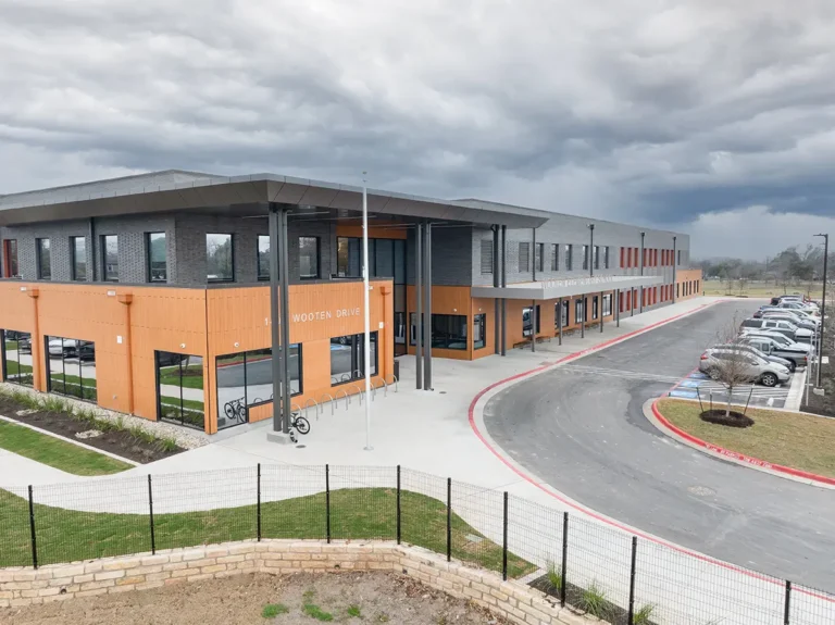 Modern two-story building with orange and gray exterior under a cloudy sky. Curved driveway and parked cars on the right, creating an organized, sleek atmosphere.