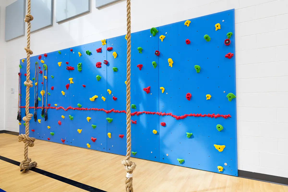 A blue indoor climbing wall with colorful holds and a horizontal red rope line. Two ropes hang from the ceiling, set against a bright gym floor.