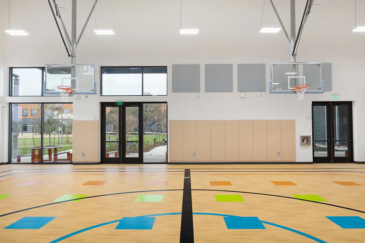 Indoor basketball court with wood flooring, colorful squares, and two hoops. Large windows and doors reveal an outdoor park. Bright, modern atmosphere.