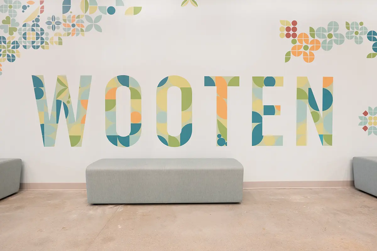 Colorful letters spelling "WOOTEN" on a wall, surrounded by geometric floral motifs. Gray benches are placed beneath, creating a modern, welcoming vibe.