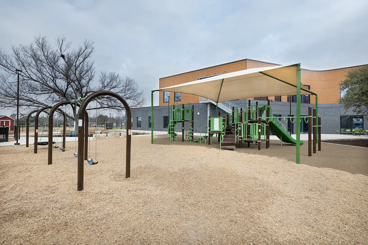 Playground with swings and green climbing structures under a sunshade. Nearby, a modern building with orange and gray tones. The atmosphere is calm.