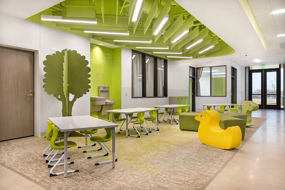Bright, modern classroom with green accents. Features include tables with green chairs, playful yellow duck seats, and tree-shaped dividers creating a lively atmosphere.