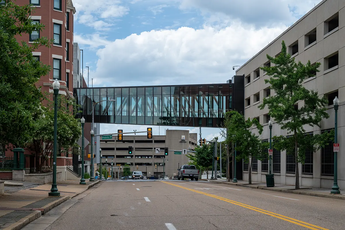 Urban street view with a glass pedestrian bridge connecting two buildings. Trees line the road, and cars are stopped at a traffic light under a cloudy sky.