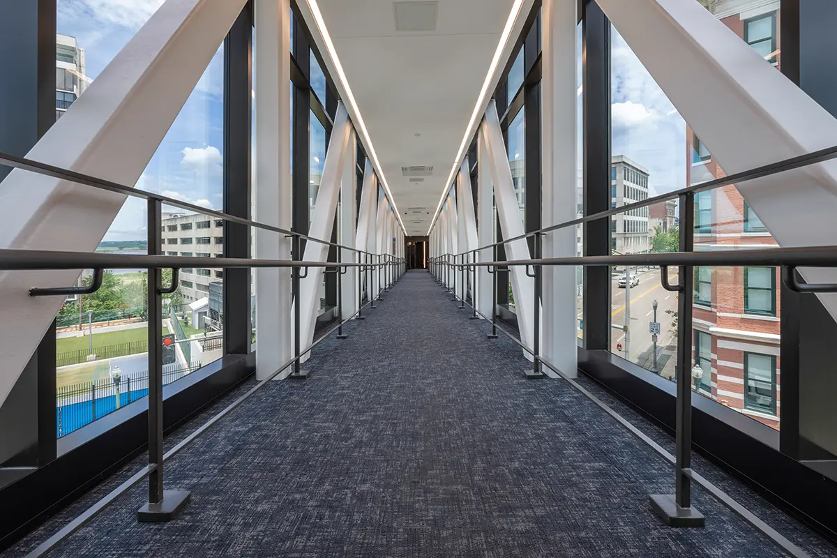 A long, glass-walled pedestrian bridge with carpeted flooring. Metal railings line the sides, and sunlight streams in, creating a bright, open feel.