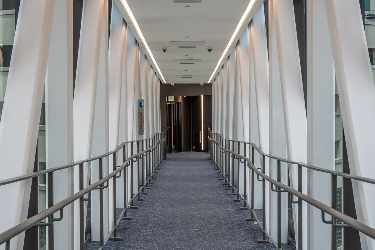 Modern glass-enclosed walkway with angled supports and metal railings, leading to double doors. Bright lighting and clean design evoke a futuristic feel.