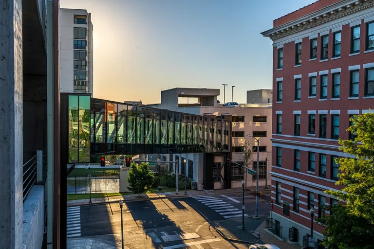 Urban scene with a glass pedestrian bridge connecting buildings over a city street. The sun sets in the background, casting long shadows.