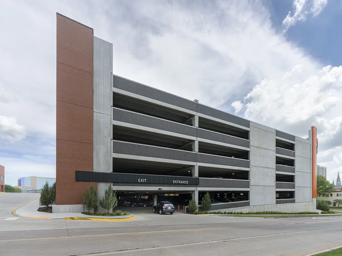 Five-story parking garage with beige and red brick exterior under a partly cloudy sky. Entrance and exit signs are visible, with a car entering.