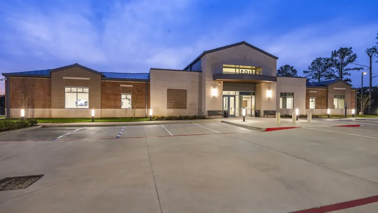 Small police station at dusk, featuring a modern design with brick and beige walls. The building is illuminated, and an empty parking lot surrounds it.