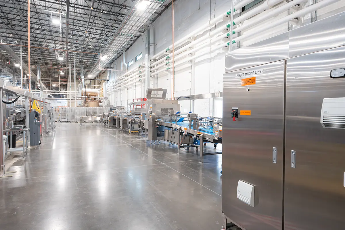 A large, bright factory interior with shiny metal machinery and conveyor belts. The floor is polished, and overhead pipes line the high ceiling.