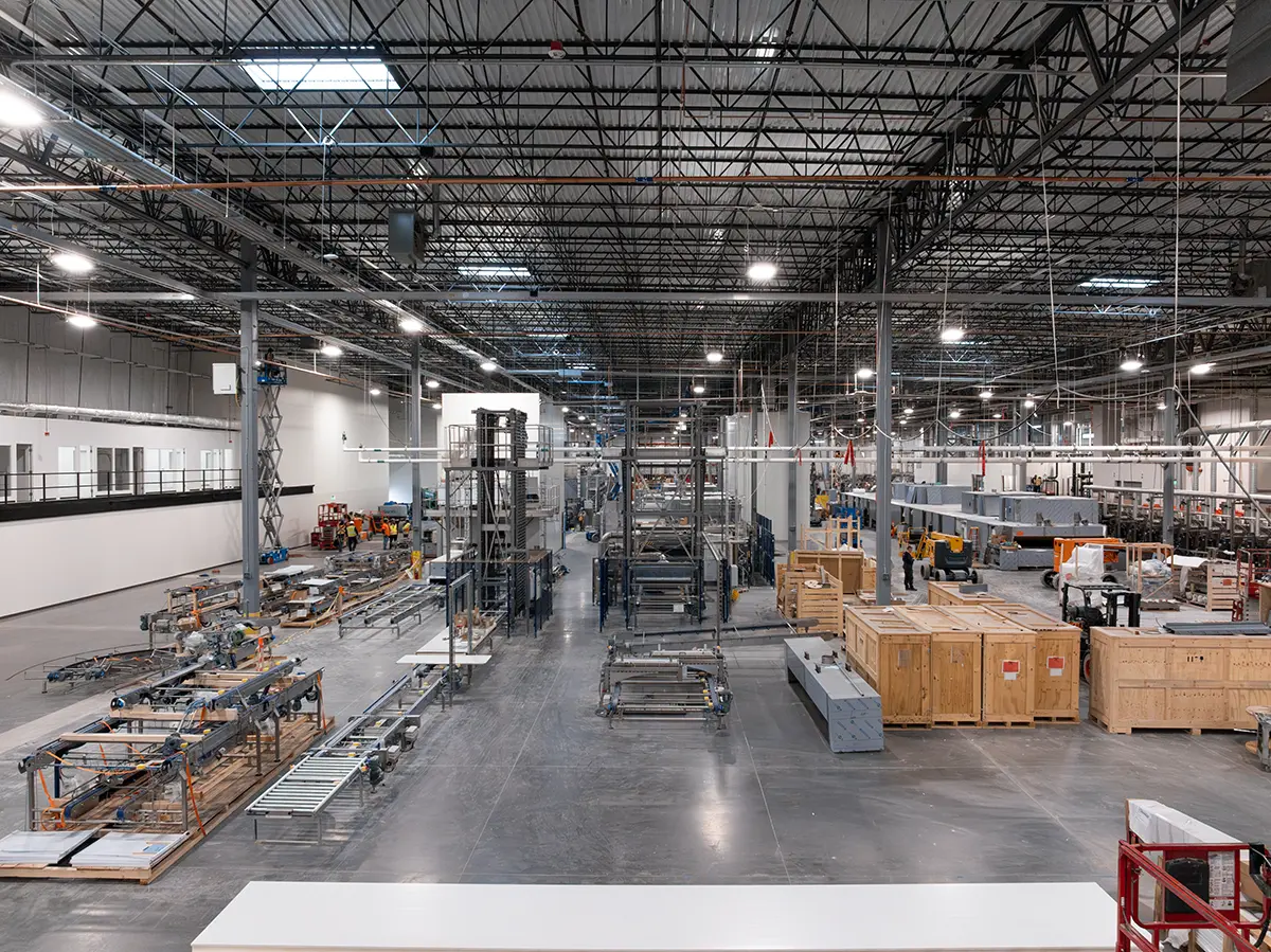 Spacious industrial warehouse with metal framework, machinery, and wooden crates. The bright lighting emphasizes the orderly and busy atmosphere.