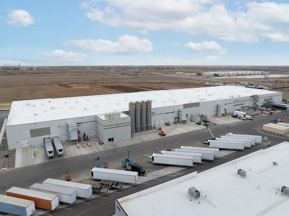 Aerial view of a large industrial facility with a white roof, surrounded by barren land. Multiple trailers are parked outside, conveying a busy and functional atmosphere.