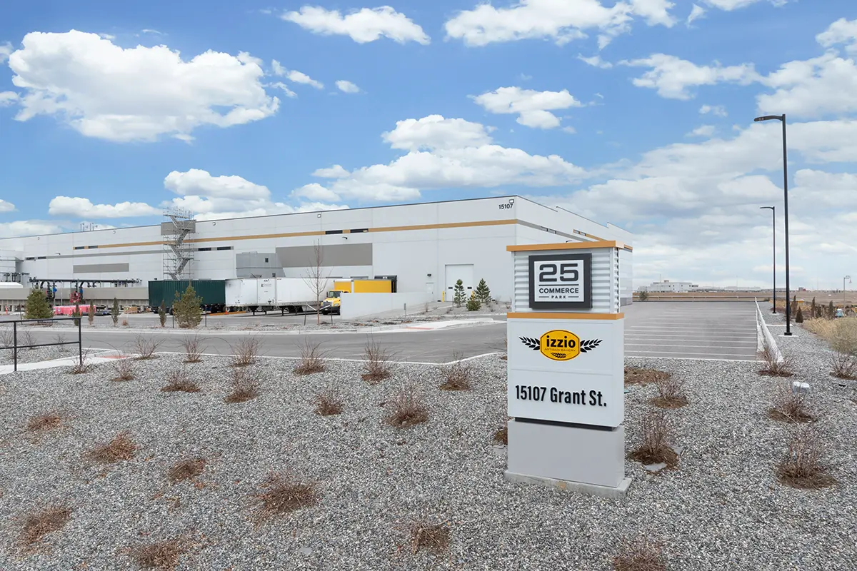 Industrial building with trucks and loading docks, set against a blue sky with clouds. Foreground shows a sign reading "25 Commerce, Izzio, 15107 Grant St." Sparse shrubs decorate the rocky landscape, creating a calm, industrial tone.