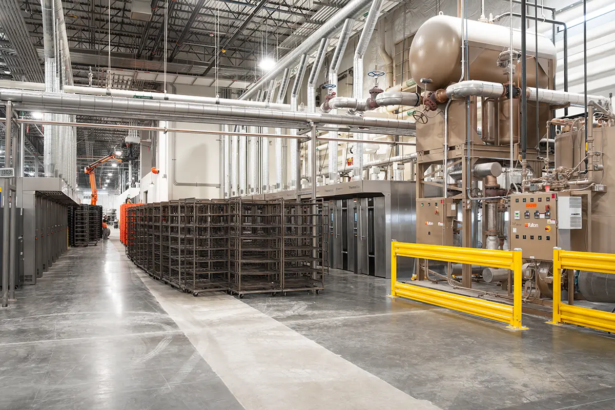 Industrial interior with metal machinery, pipework, and empty racks in a spacious warehouse. Bright lights and a high ceiling convey efficiency.