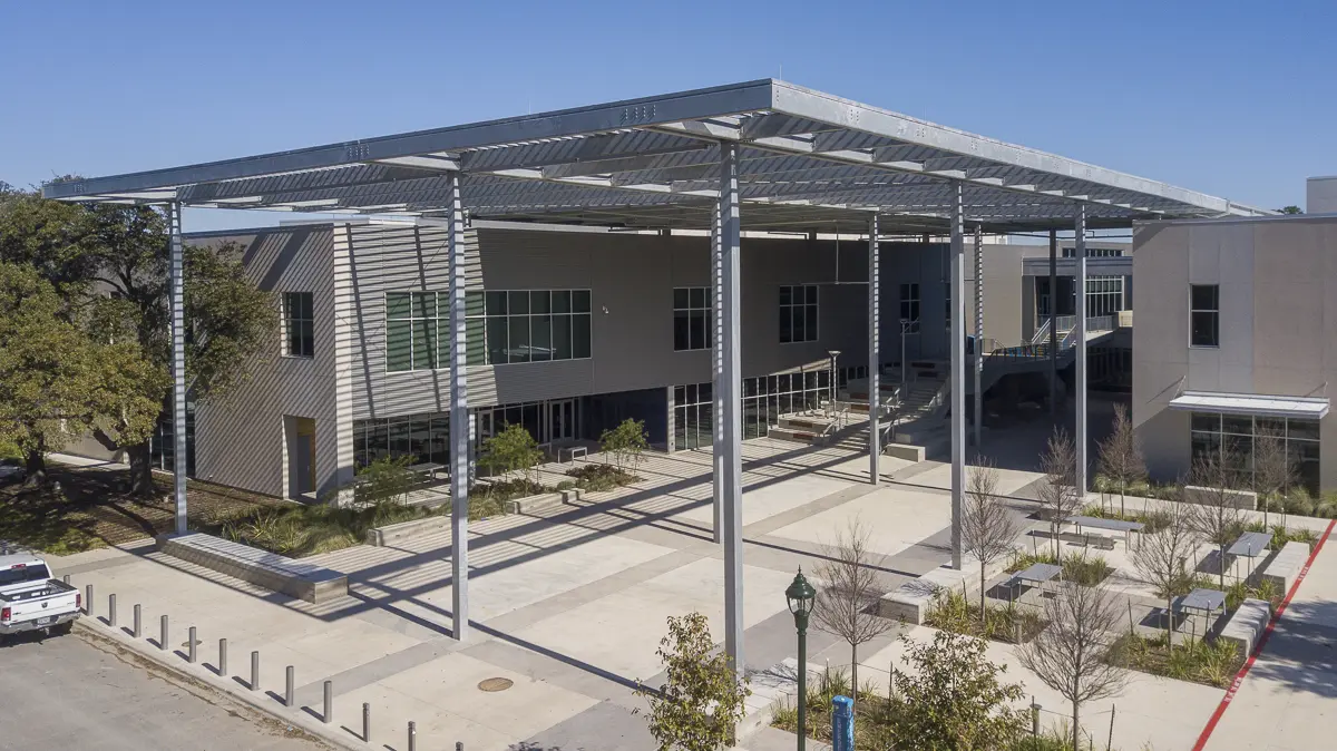 Modern building with a large metal canopy, featuring glass windows and beige walls. Surrounding landscape includes trees and paved walkways, under a clear blue sky.