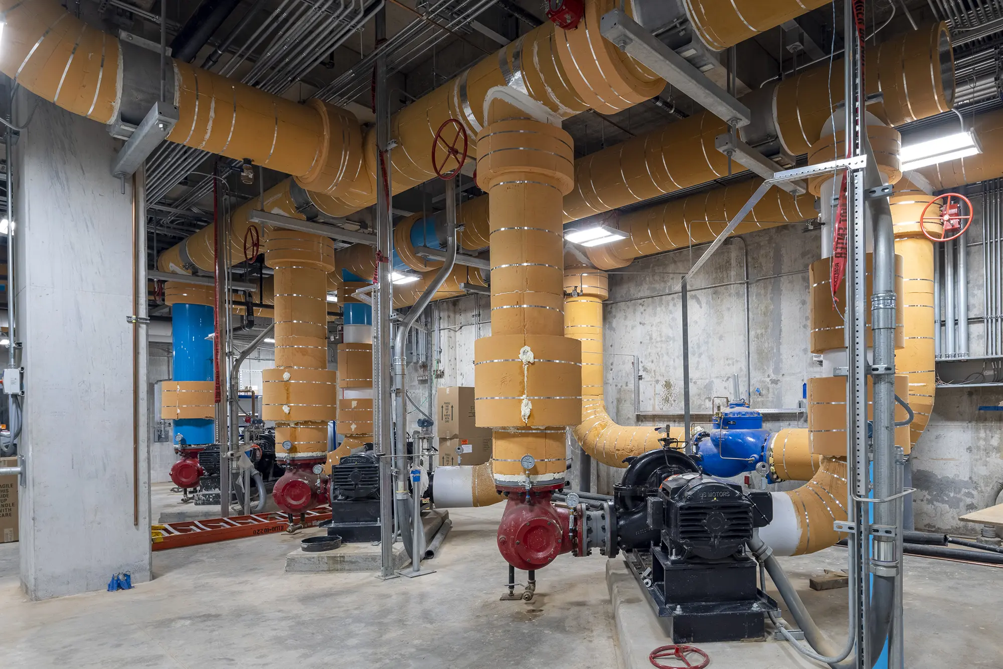 Utility room with large orange insulated pipes, red valves, and black industrial pumps. The concrete walls and exposed ceilings give an industrial feel.
