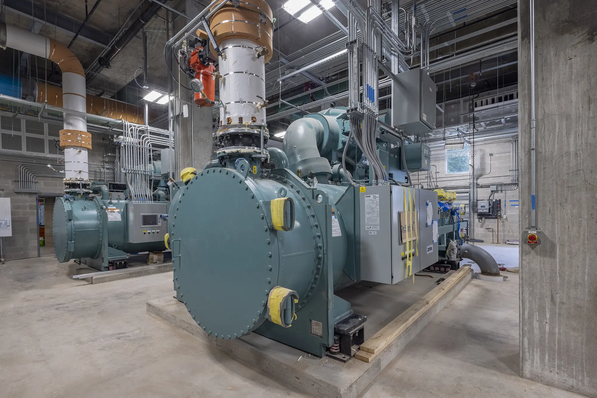 Interior of a large industrial facility with several massive, green machines featuring pipes and cables, situated on a concrete floor, under bright lights.