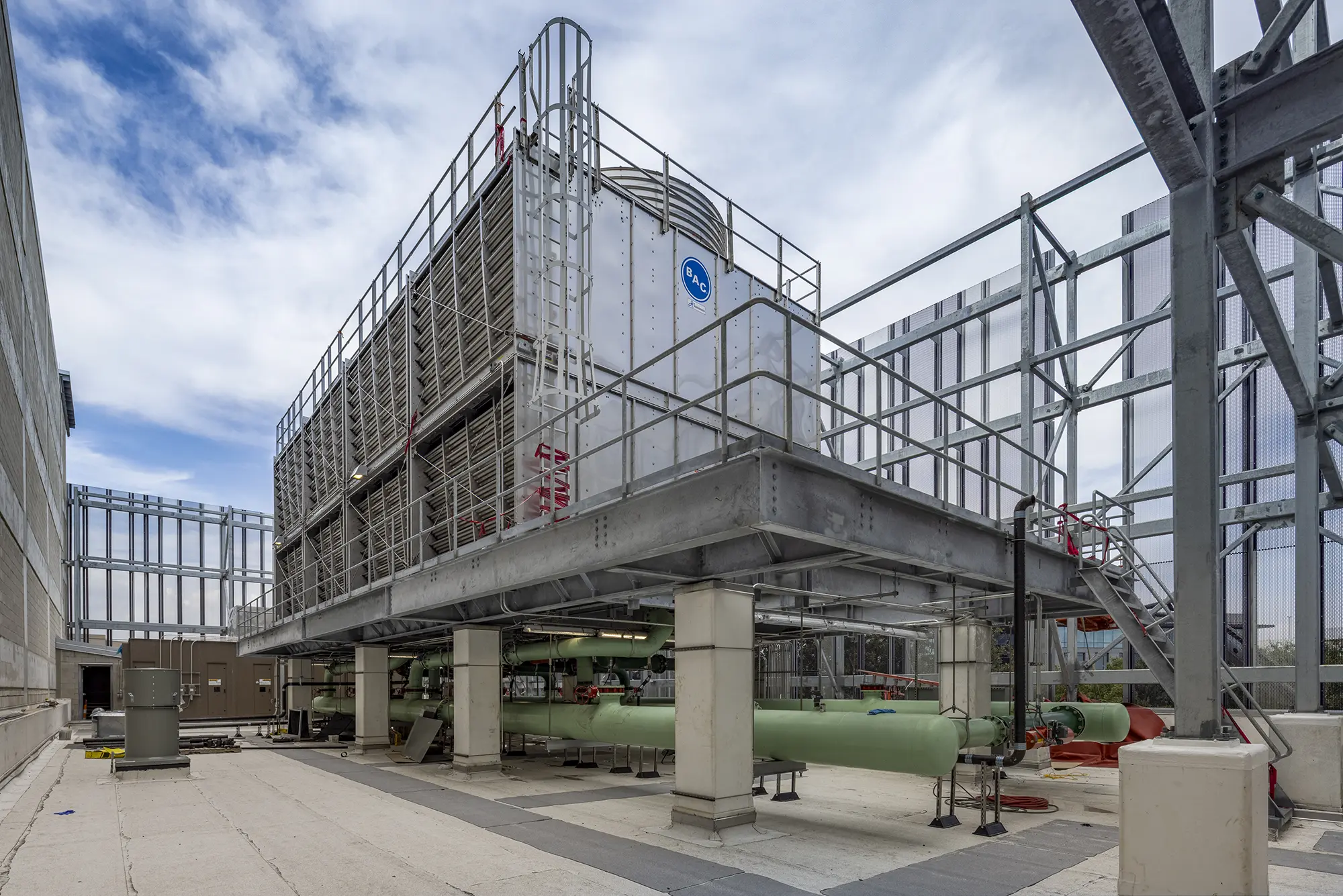 Industrial cooling tower on rooftop, elevated on concrete pillars. Surrounded by metal railings and pipes. Sky is partly cloudy, creating a neutral tone.