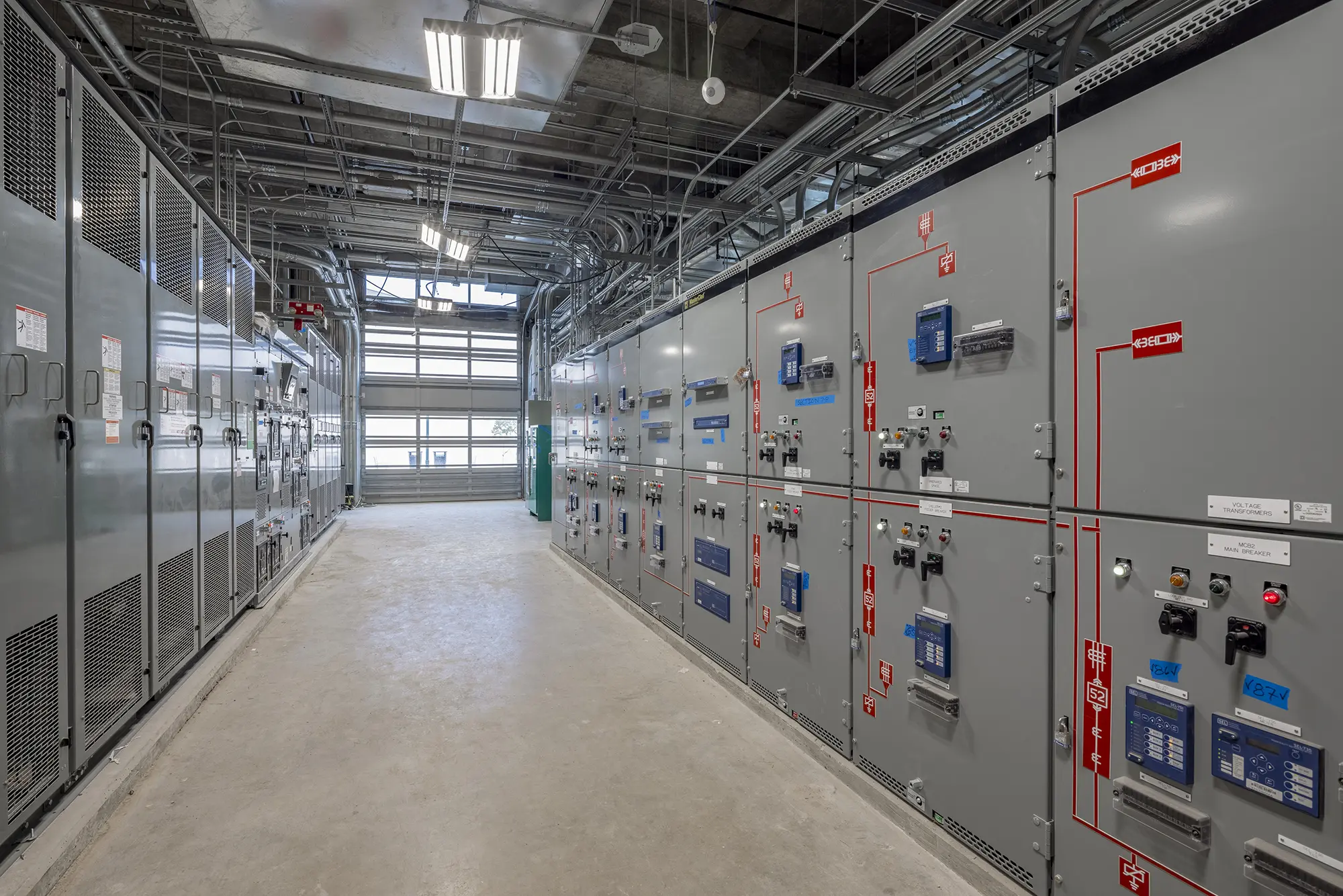 Corridor with electrical control panels lining the walls, featuring various switches and labels. Overhead lighting and visible wiring create an industrial tone.
