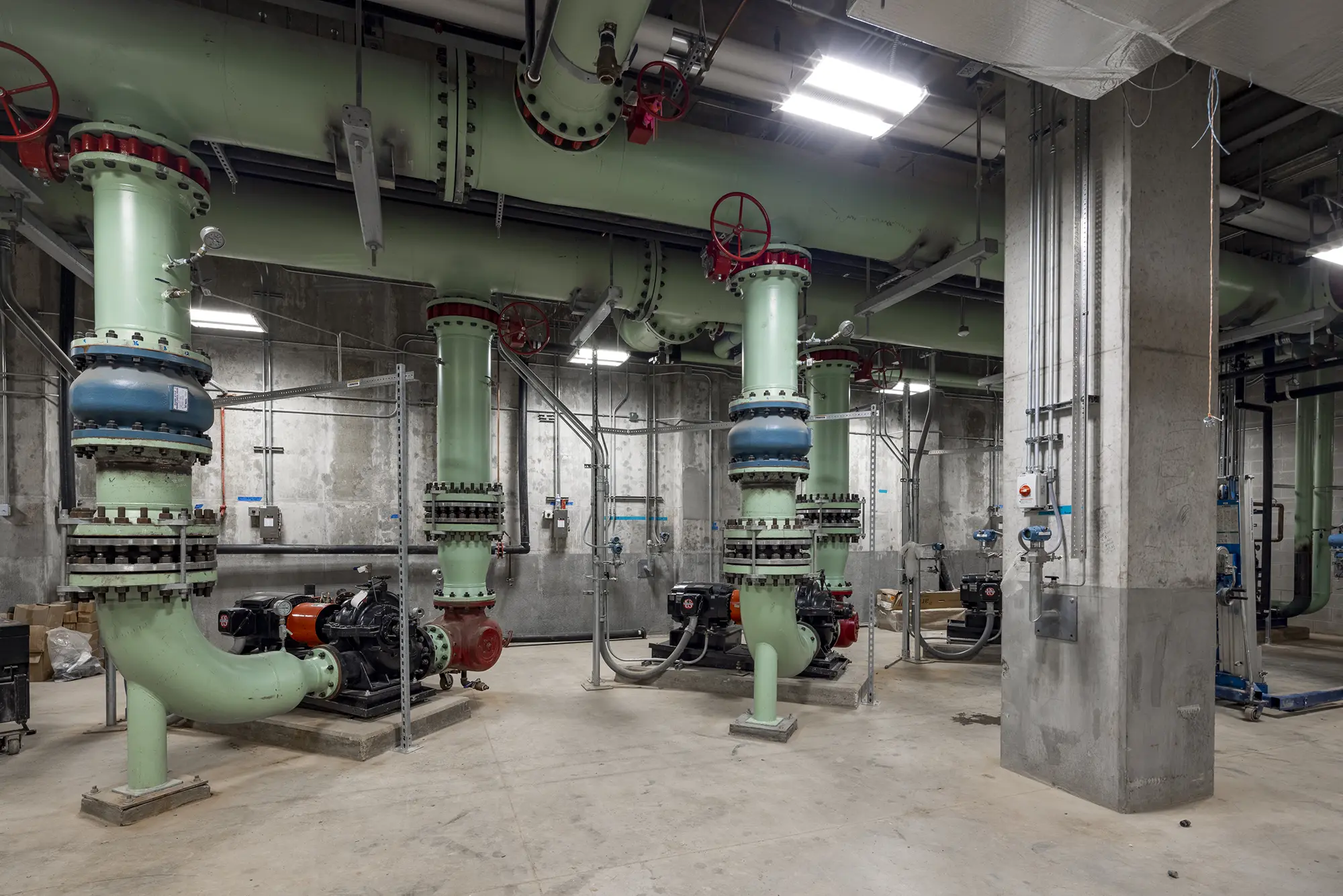 A large, industrial room with multiple pastel green pipes and red valves. The concrete walls and floor add a utilitarian feel. Bright overhead lights illuminate the space.