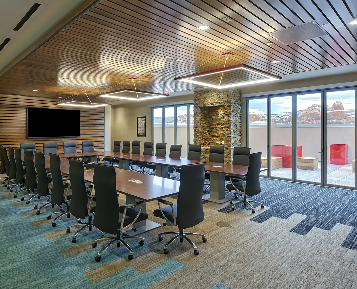 Modern conference room with a U-shaped wooden table, black swivel chairs, large windows revealing a scenic mountain view. Warm lighting and stone wall accentuate the space.