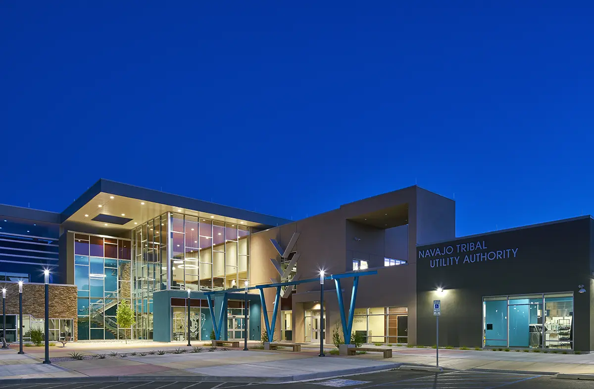 Modern building at dusk labeled "Navajo Tribal Utility Authority," illuminated with vibrant lights, showcasing large glass windows and contemporary design.