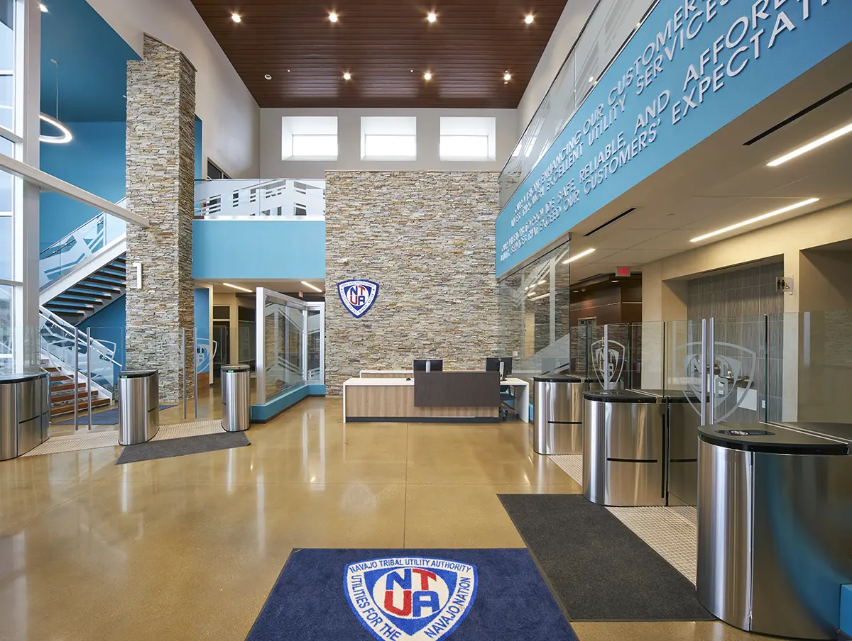 Large, modern lobby with high ceilings and natural light. Features stone walls, blue accents, turnstiles, and a blue mat with NTUA logo. Inviting and professional.
