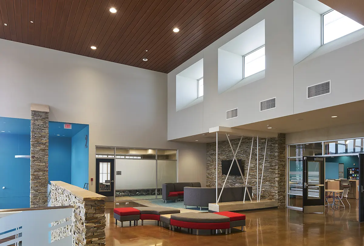 Large, modern lobby with high ceilings and natural light. Features stone walls, blue accents, turnstiles, and a blue mat with NTUA logo. Inviting and professional.
