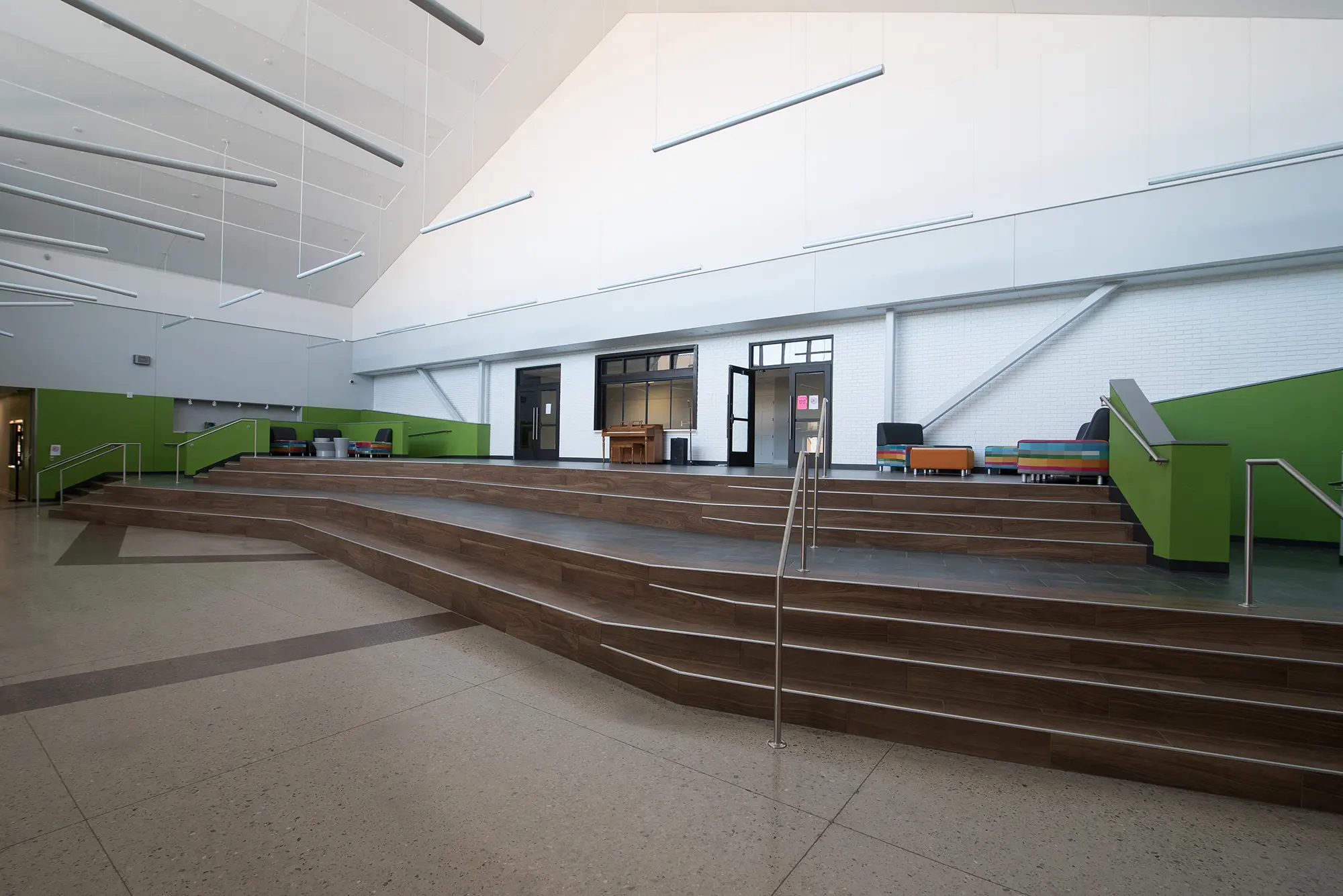 Spacious modern interior with tiered wooden seating and green accent walls. High ceiling, large glass doors, and scattered colorful chairs evoke a calm, open ambiance.