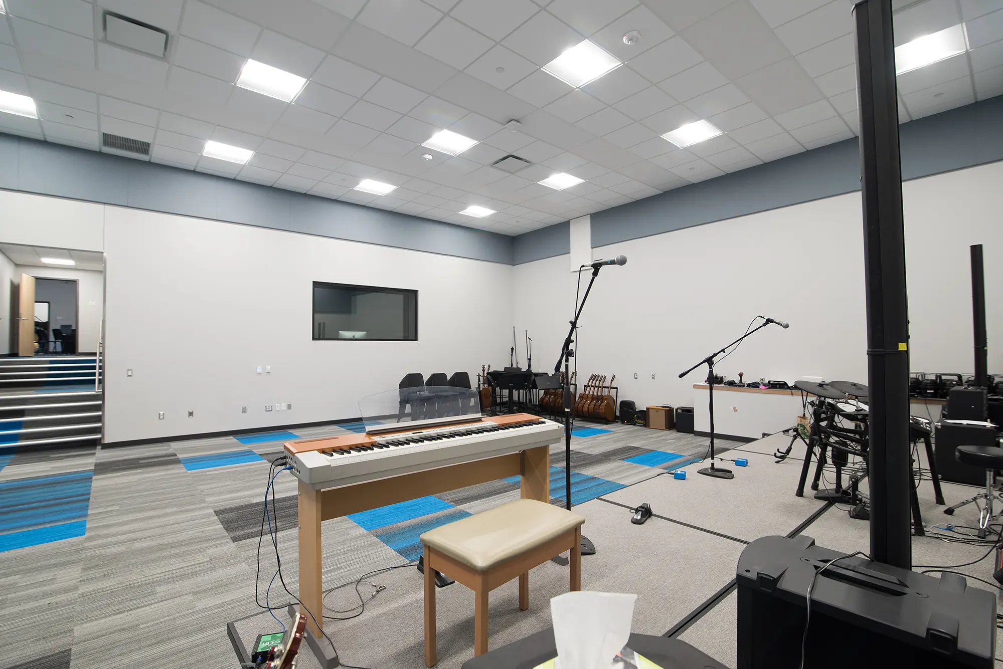 Modern music rehearsal room with a keyboard, stool, and multiple microphones. The room has blue and gray carpeting, white walls, and a window. Calm atmosphere.