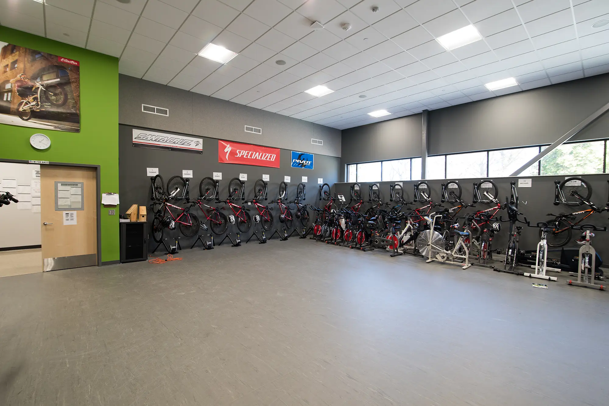 A spacious room with gray walls and ceiling holds lines of stationary bicycles mounted on racks. Banners of cycling brands hang above, suggesting a bike training facility.