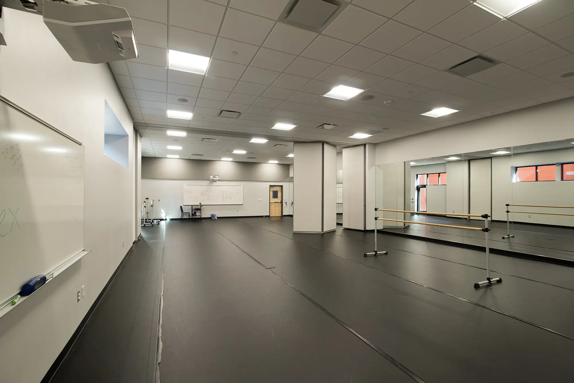 Spacious dance studio with black flooring, mirrored wall, and ballet barres. Bright overhead lights and whiteboards create a clean, modern look.