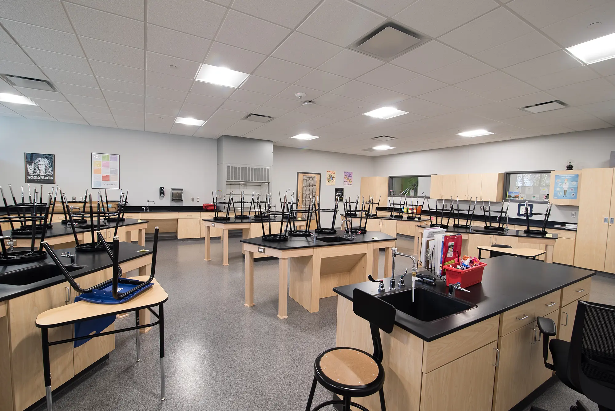 Empty science classroom with beige counters and black surfaces. Stools are placed upside down on desks. Bright lighting and educational posters decorate the walls.