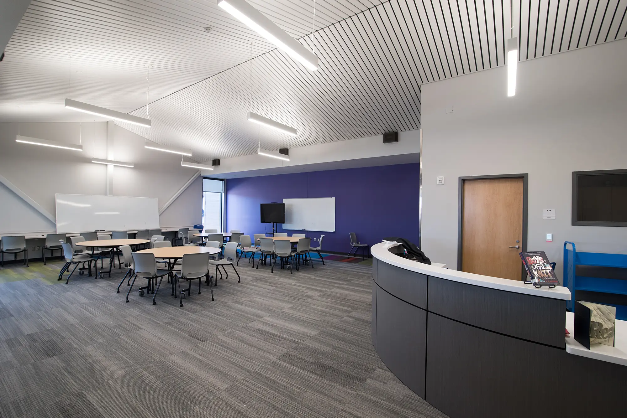 Modern classroom with round tables and chairs, a purple accent wall with a TV, whiteboards, and sleek lighting. The room feels open and collaborative.