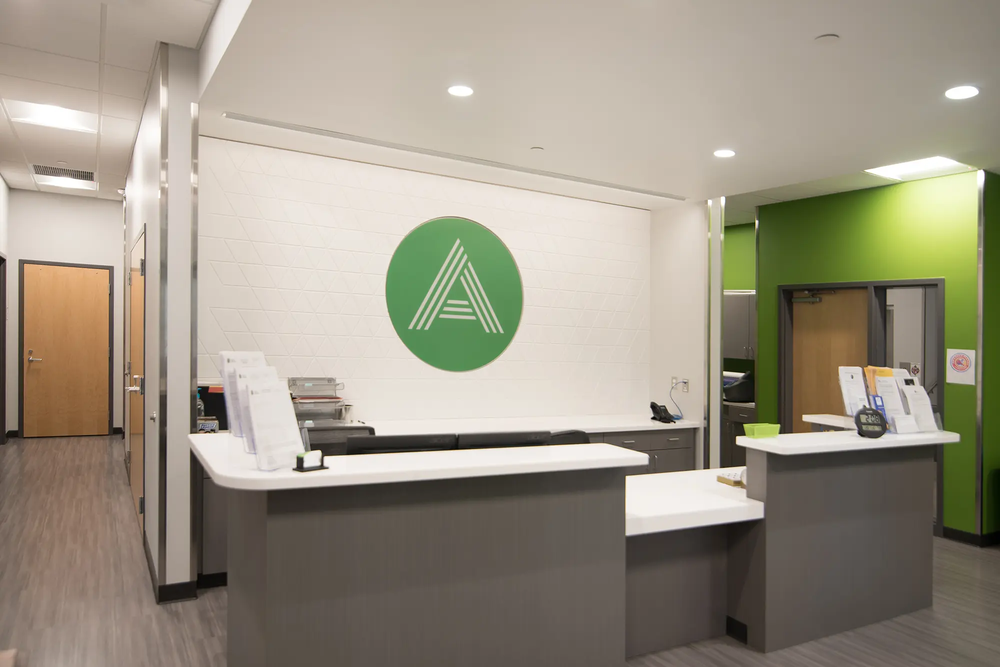 Modern reception area with gray desks and white countertops. A green circle with a stylized 'A' logo is on the wall. Bright and welcoming atmosphere.